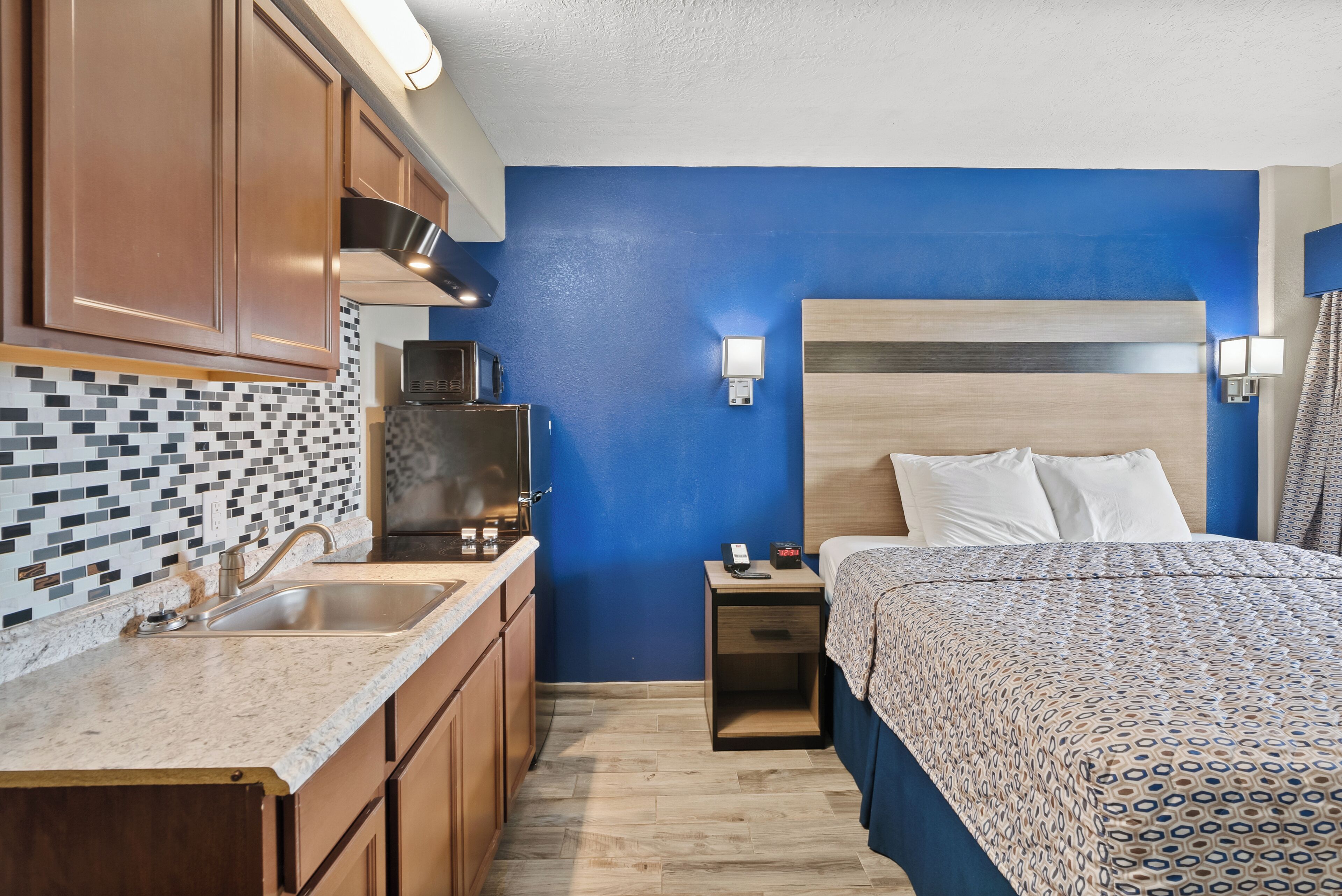 A hotel room with a blue accent wall, a wooden headboard, a bed with patterned bedding, a nightstand with a lamp, and a kitchen area with a tiled backsplash and wooden cabinets.