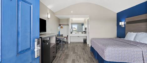 Habitación de hotel pequeña con una puerta azul, una cama con colcha estampada, una kitchenette compacta, un escritorio con silla y un espejo sobre el lavabo.