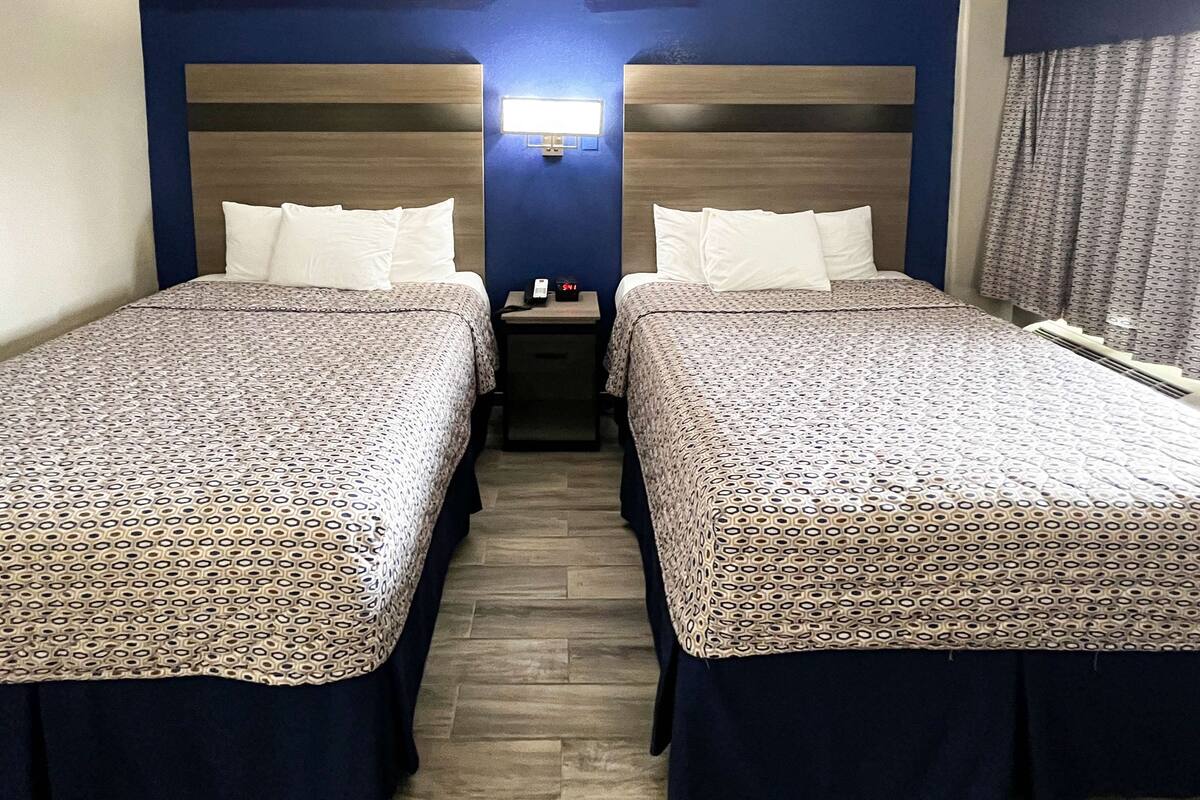 Habitación de hotel con dos camas, cada una con una colcha estampada y almohadas a juego. Entre las camas, un mesita de noche con un reloj y un teléfono. La habitación tiene un cabecero de madera y una pared decorativa azul.