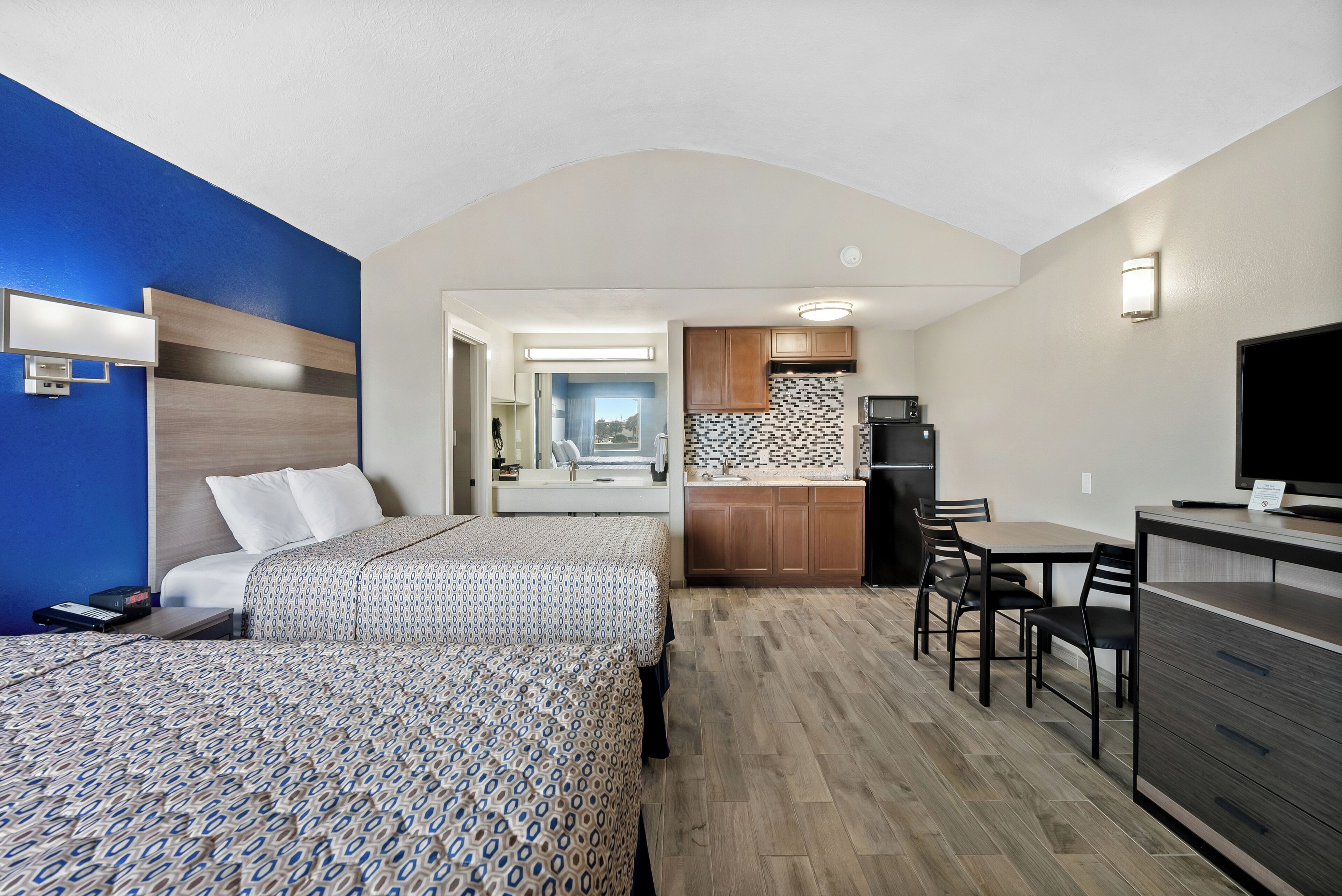 A hotel room with two beds, a kitchenette, a dining area, and a bathroom visible through an open door.