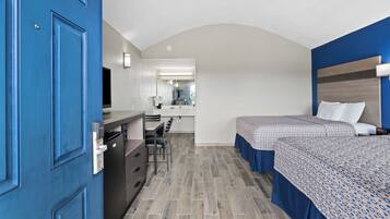 Une chambre d’hôtel avec une porte bleue, un bureau avec une chaise, un lit recouvert d’une couvre-lit à motifs, et une salle de bain visible au fond.