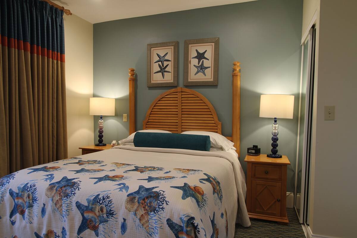 Une chambre d’hôtel avec un lit, deux étoiles de mer décoratives fixées au mur, une tête de lit en bois, deux tables de chevet avec des lampes et une fenêtre avec des rideaux.