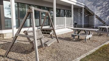 Un conjunto de columpios de madera y mesas de picnic frente a un edificio con ventanas grandes y un porche cubierto.