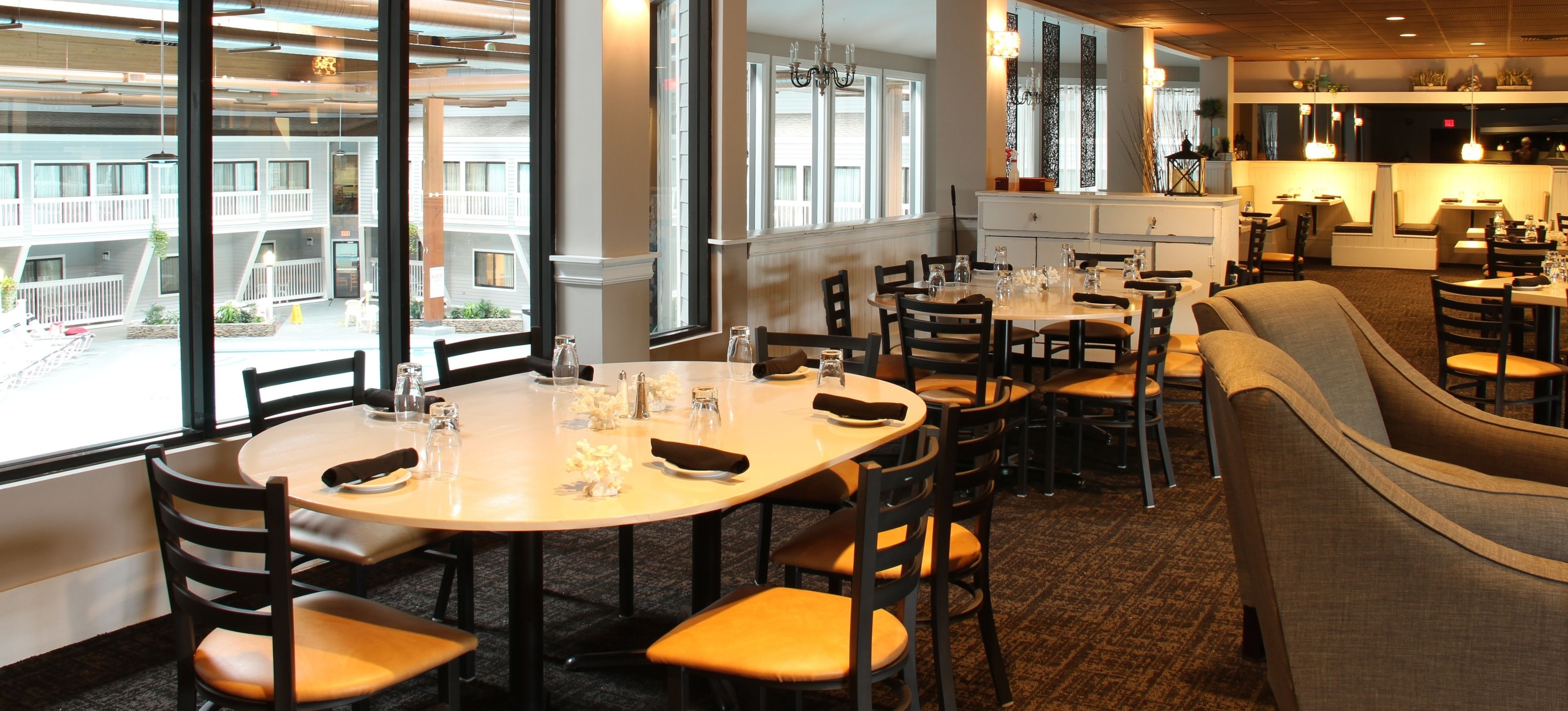 A dining area with round tables and chairs, large windows with a view of a building, and a kitchen area in the background.