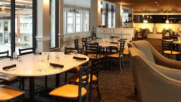 A dining area with round tables and chairs, large windows with a view of a building, and a kitchen area in the background.