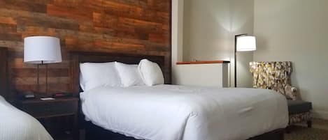 Et hotelværelse med sengegavl i træ, seng med hvide sengetøj, et natbord med lampe og en stol med mønstret pude.
