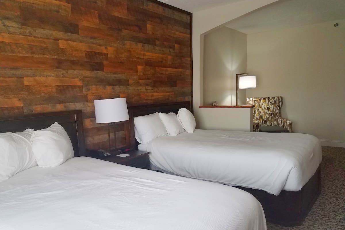 A hotel room with two beds, a wooden accent wall, a nightstand with a lamp, a mirror, and a chair.