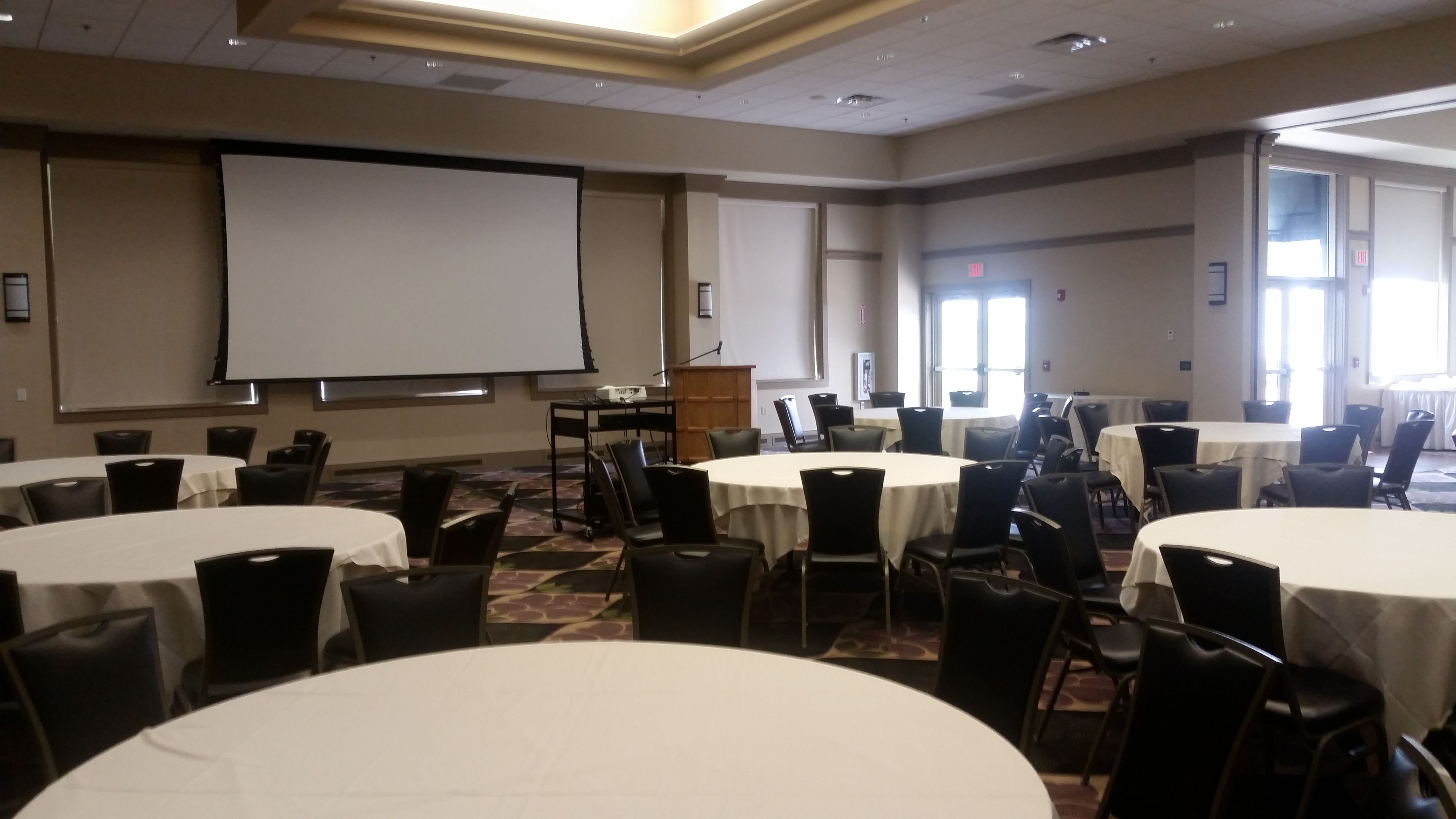 A conference room with multiple round tables, chairs, a podium, and a large projection screen.