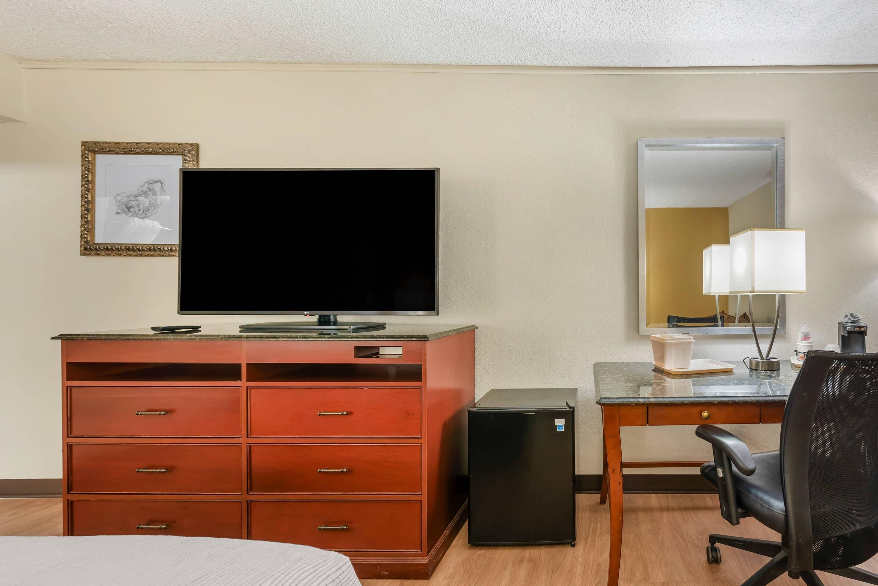 Habitación de hotel con televisión, escritorio con espejo, refrigerador pequeño y cama.