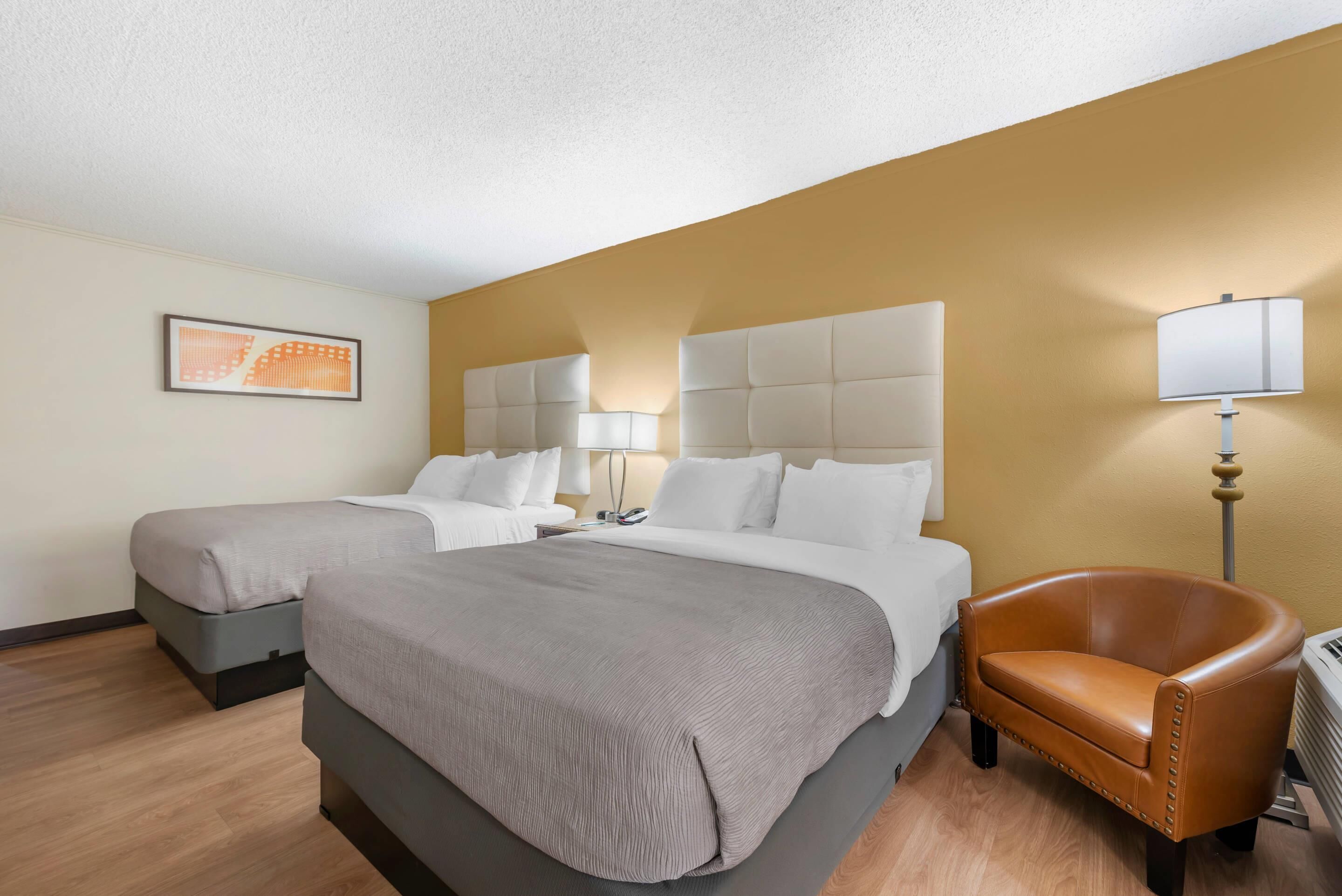 Quarto de hotel com duas camas, uma poltrona de couro marrom, uma luminária e um quadro na parede.