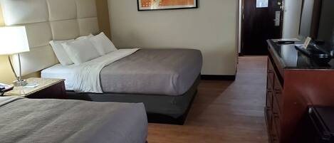 A hotel room with two beds, a desk with a lamp, a framed picture on the wall, and a dresser.