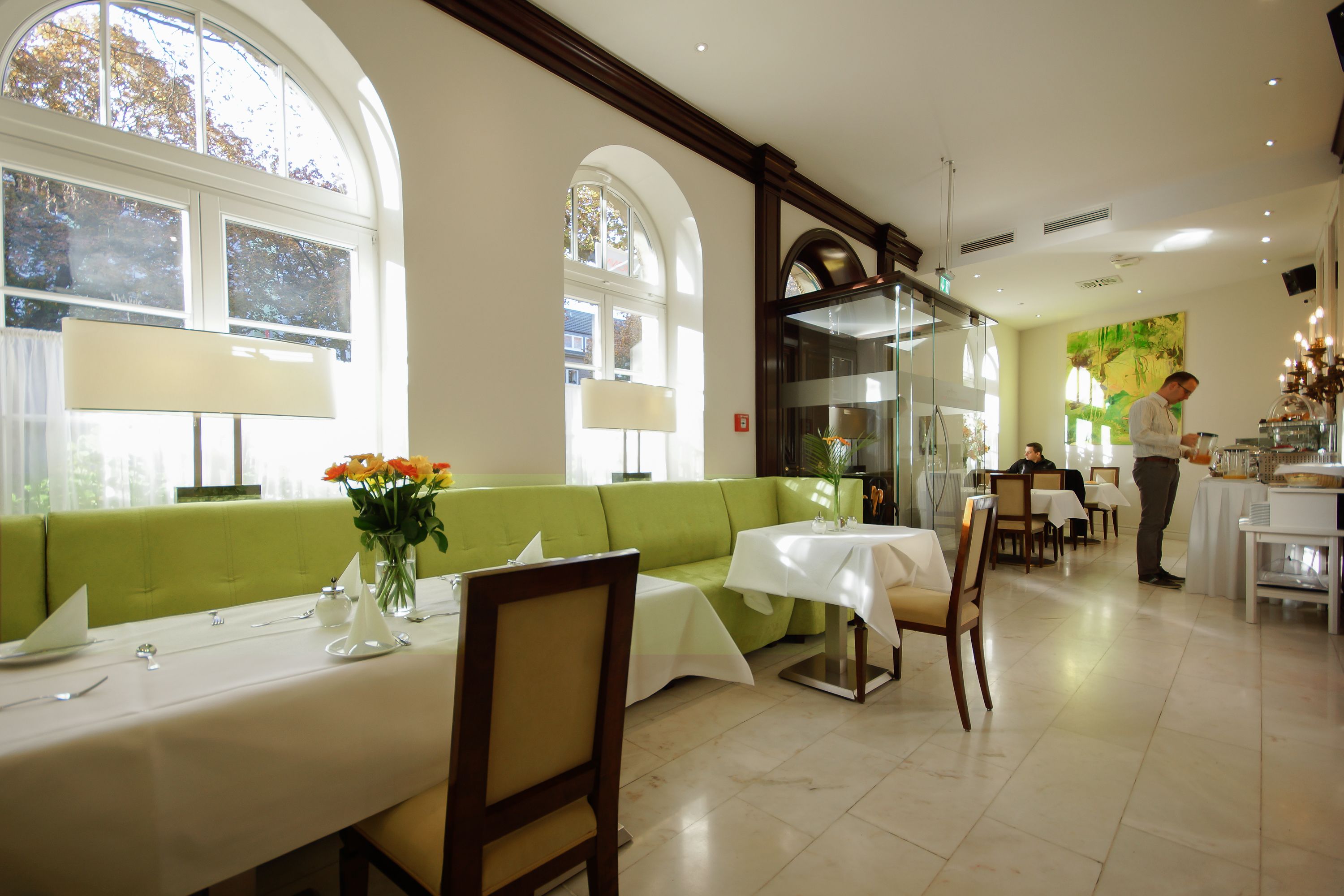 A dining area with a green sofa, tables with white tablecloths, and a waiter serving.