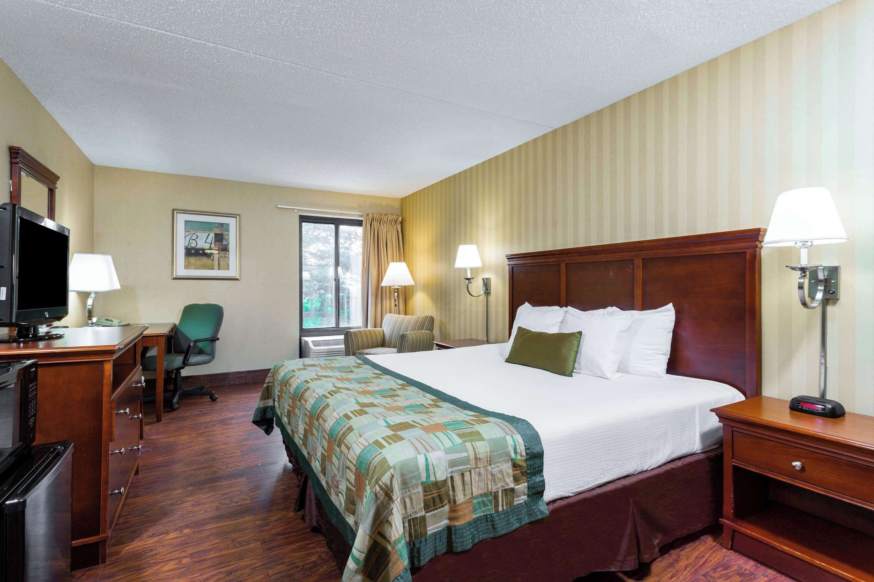 Habitación de hotel con una cama grande, un escritorio con computadora, una silla, un televisor y una ventana con cortinas.