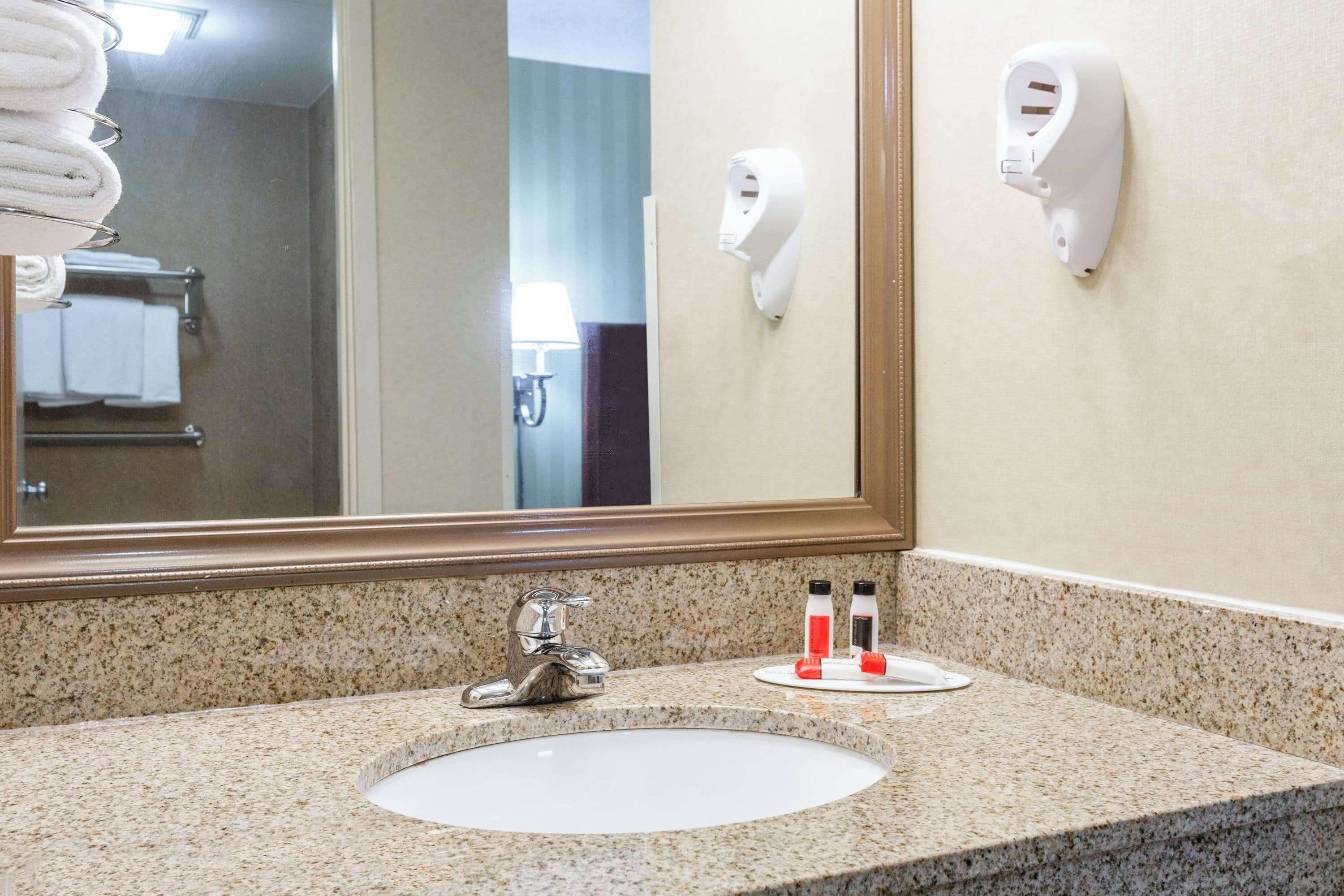Baño de hotel con lavamanos de encimera de granito, un espejo grande, un perchero con toallas dobladas y un secador de manos montado en la pared.