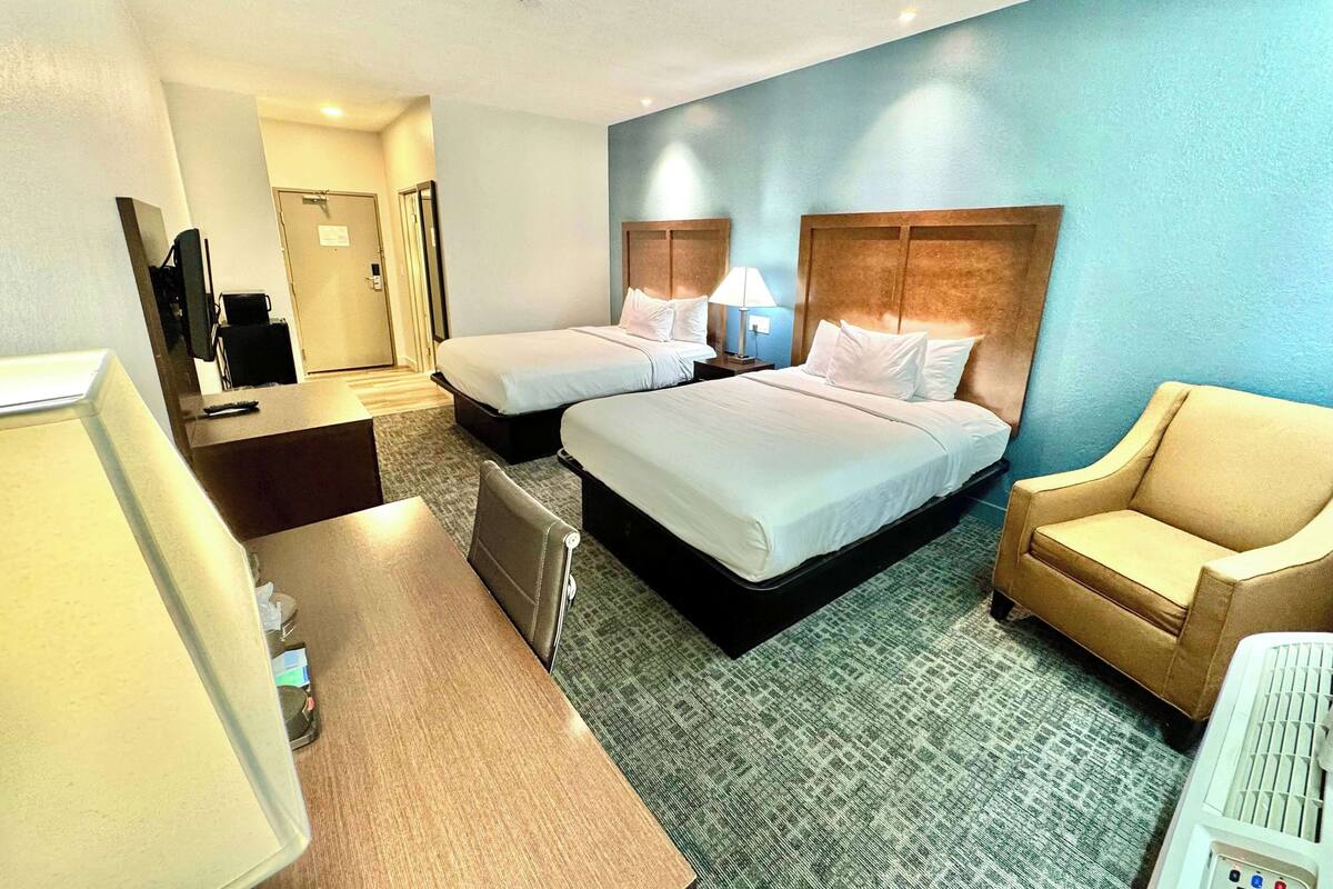 A hotel room with two beds, a desk, a chair, a TV, and a bathroom visible in the background.