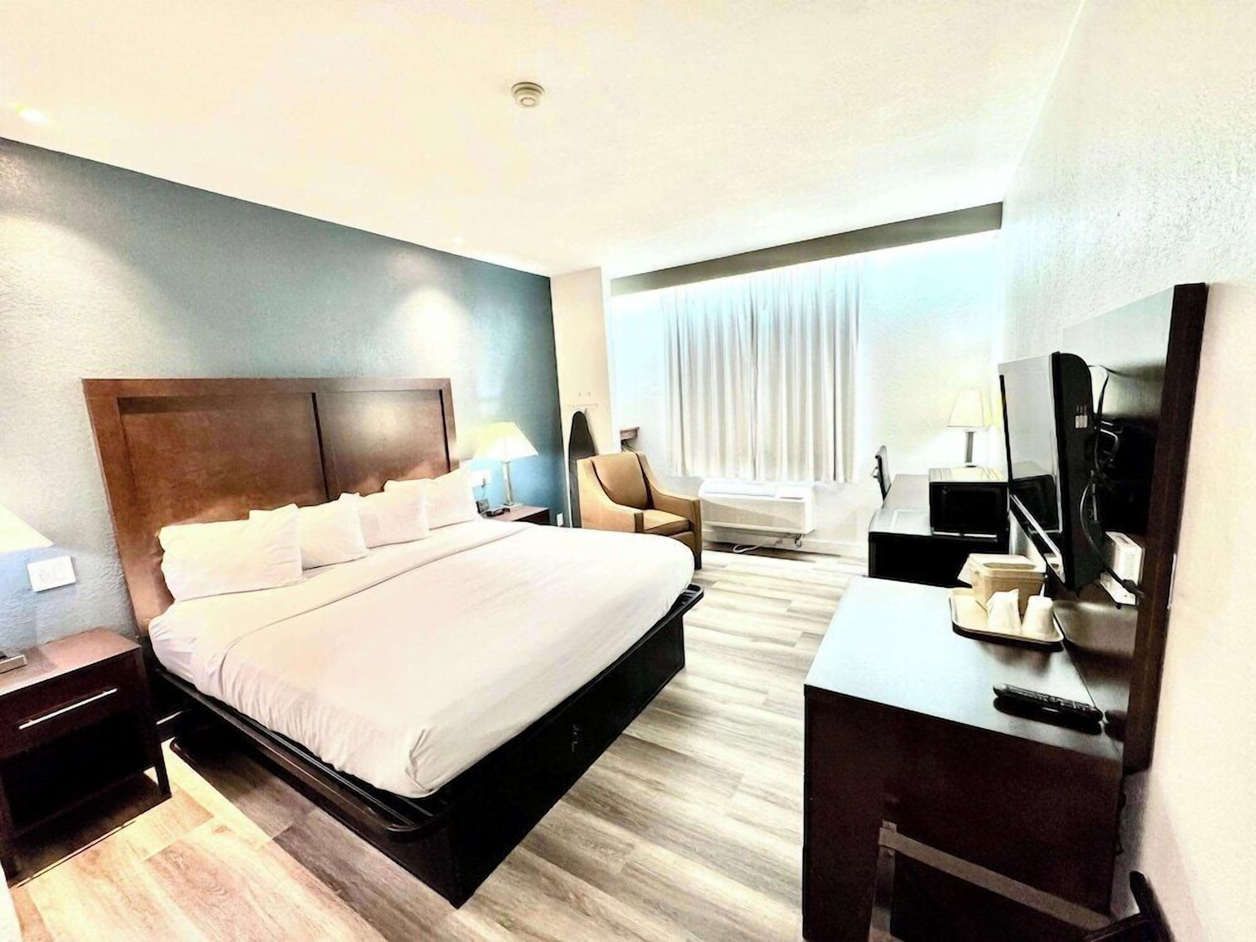 Une chambre d’hôtel avec un lit, un bureau équipé d’un ordinateur, une chaise, une télévision et une fenêtre avec des rideaux.