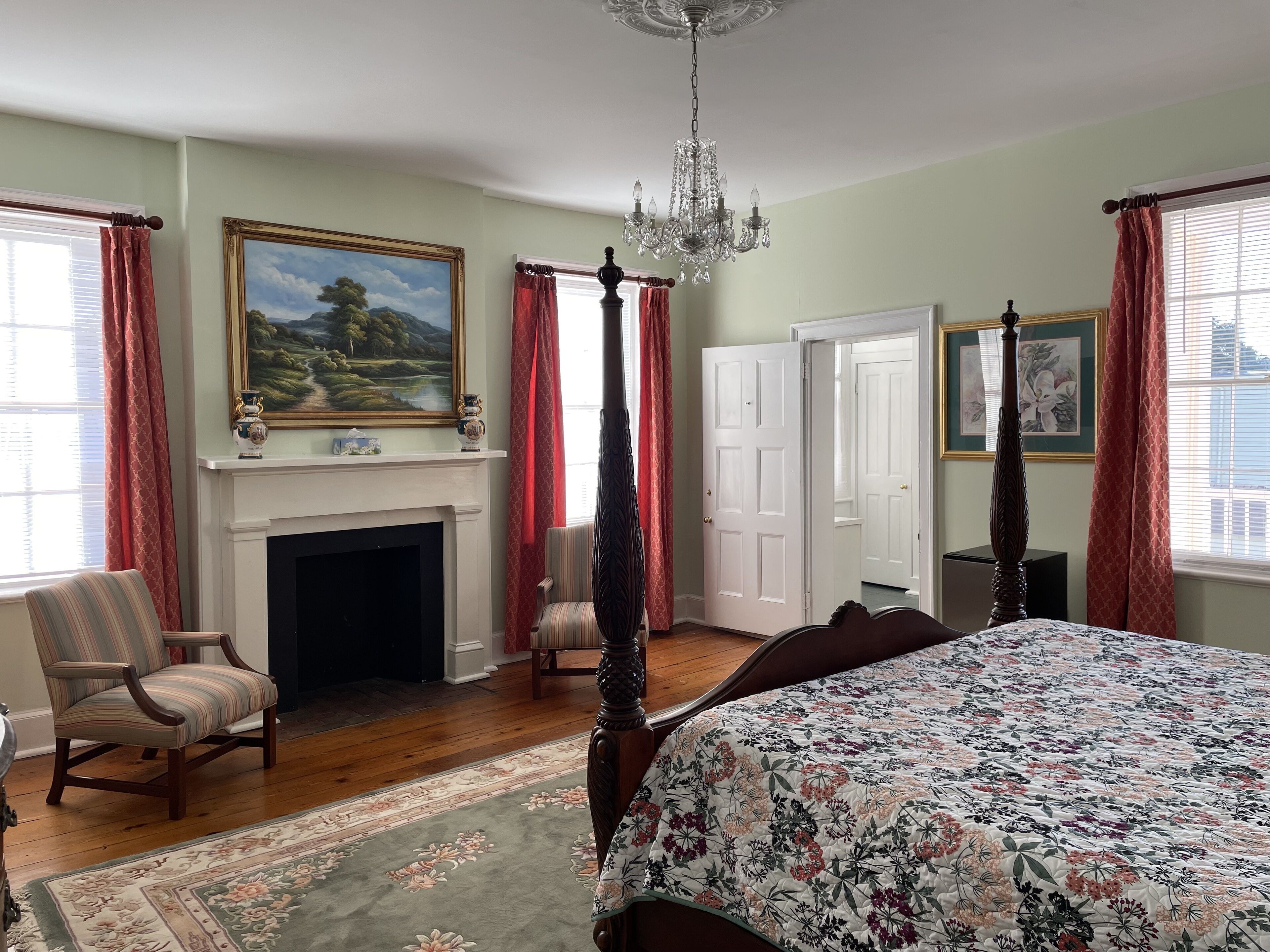A bedroom with a four-poster bed, a fireplace, a chandelier, and a large landscape painting.