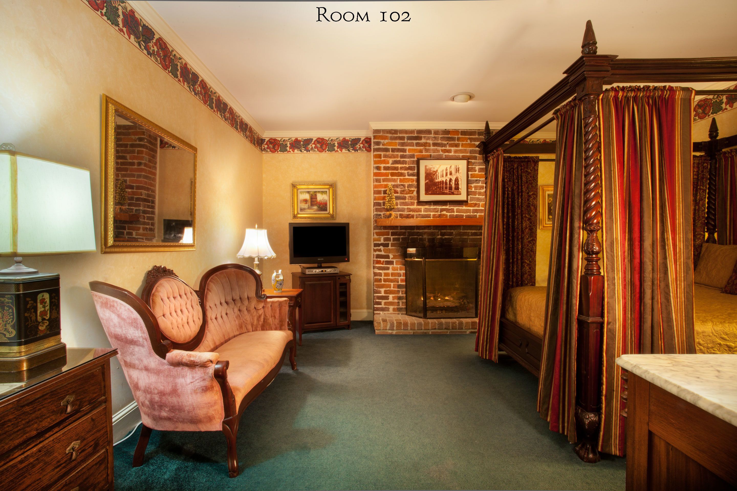 A room with a brick fireplace, a four-poster bed, a television, and a vintage armchair.