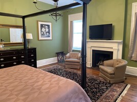A bedroom with a four-poster bed, a dresser, a television above a fireplace, and a chair.