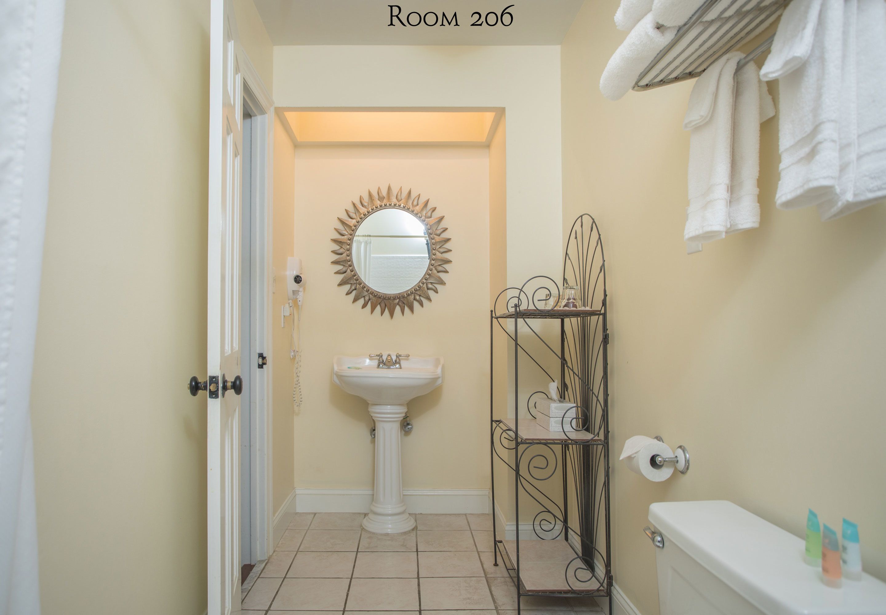 Un baño con lavamanos pedestal, espejo con diseño de sol, toallero y dispensador de papel higiénico.