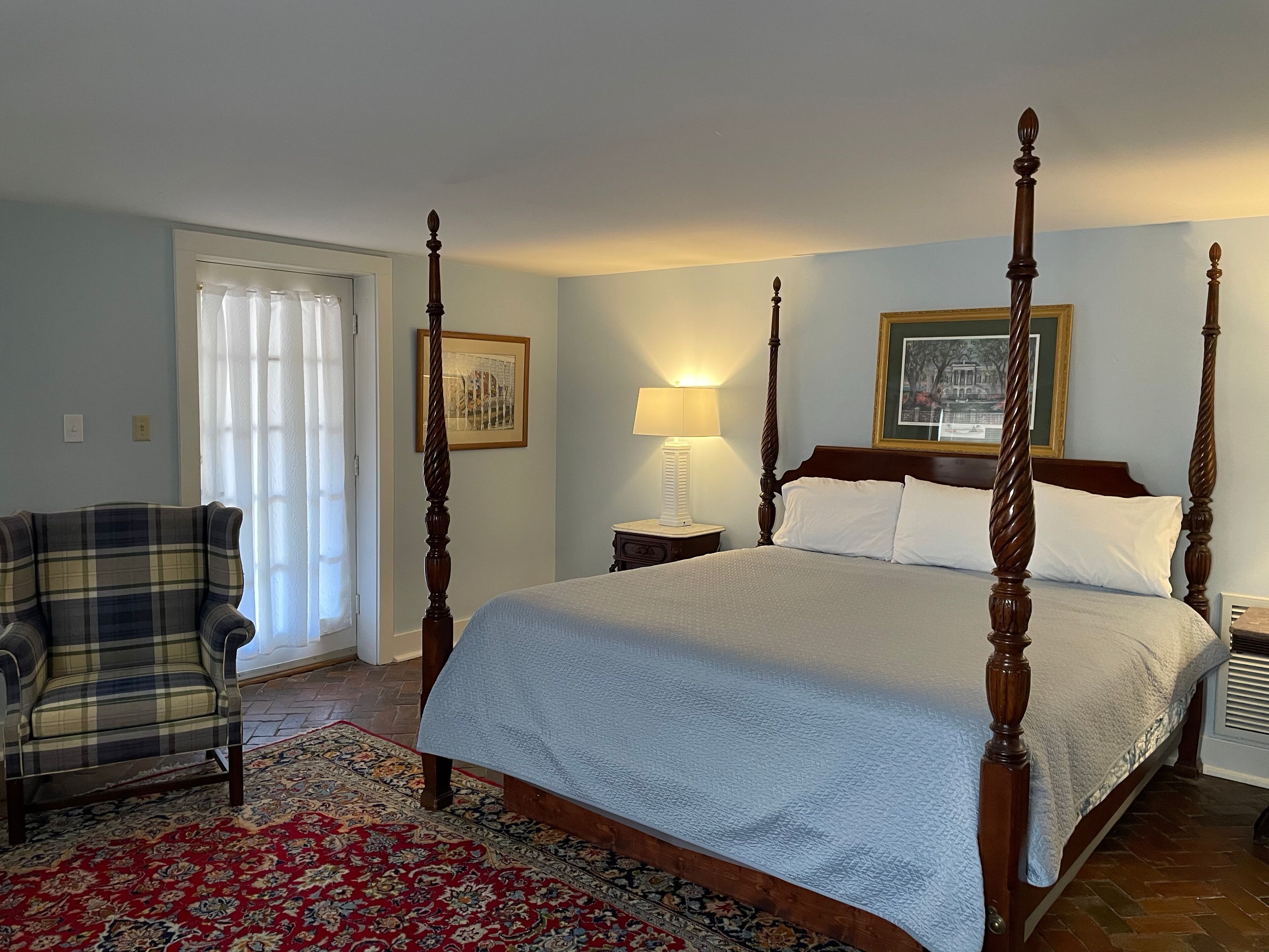 A four-poster bed with a white bedspread, a plaid armchair, a patterned rug, and a framed picture on the wall.
