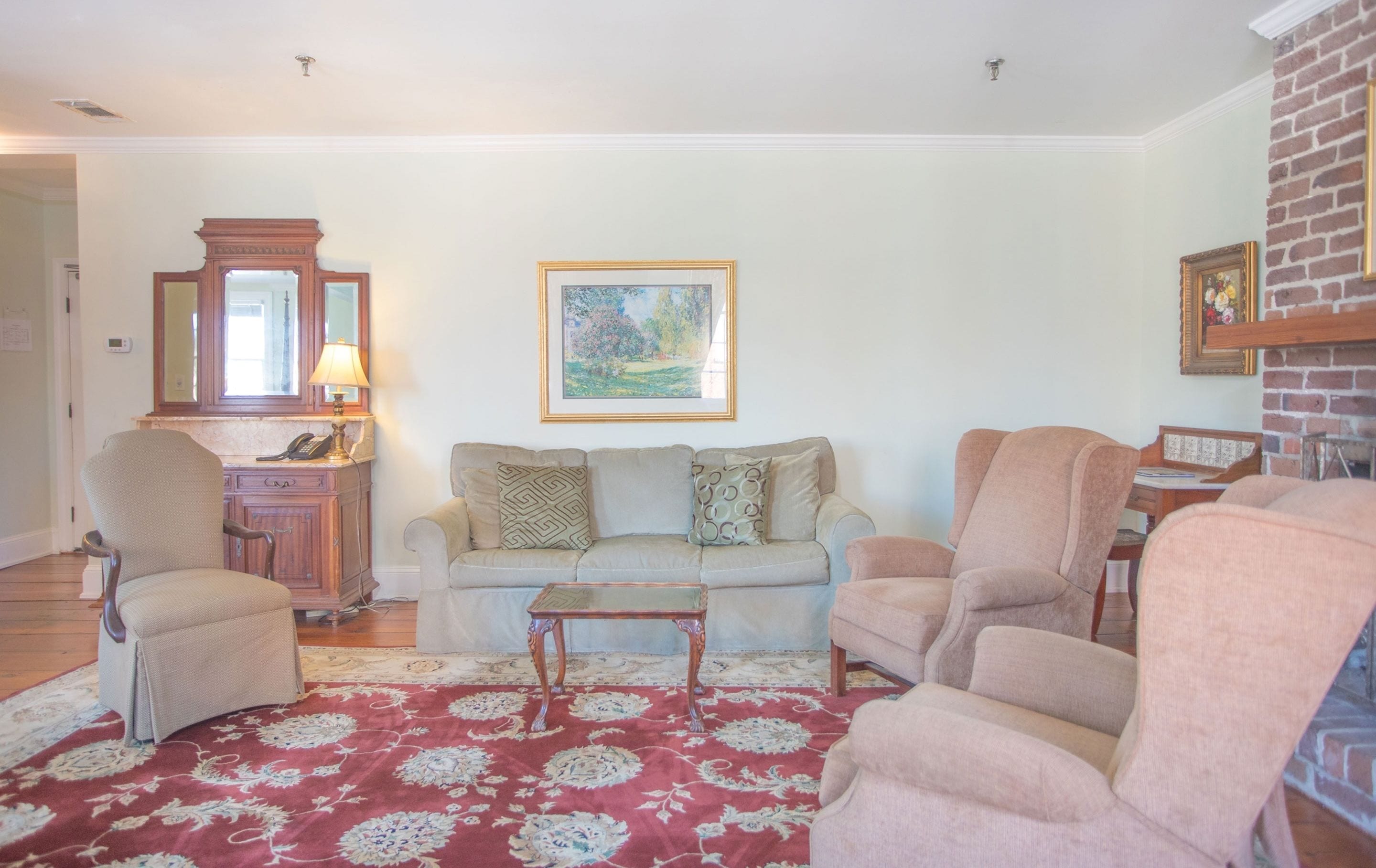 Een woonkamer met een rood kamerkleed, een beige bank, twee fauteuils, een houten bijzettafeltje en een gekaderd schilderij aan de muur.