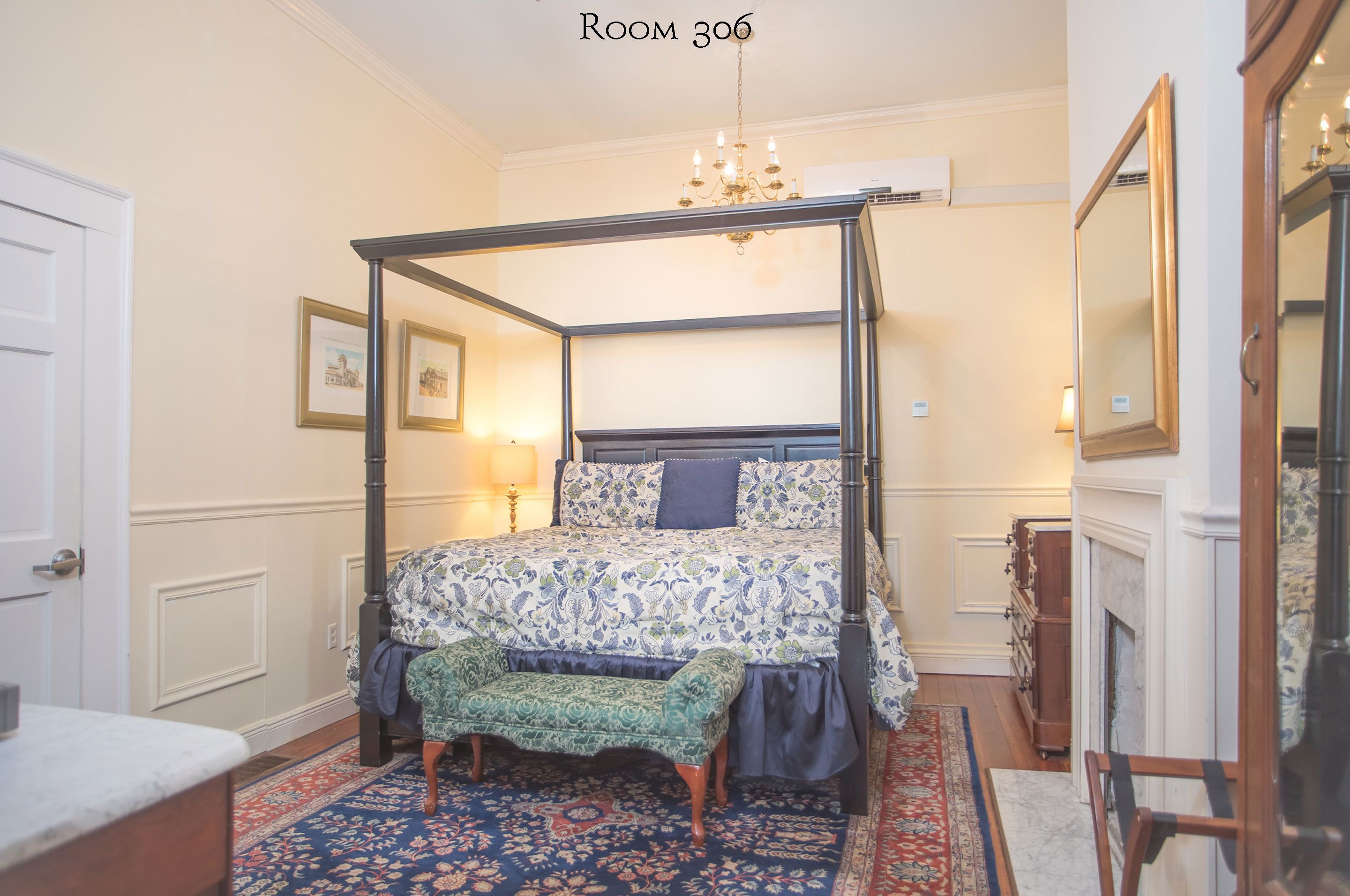 A four-poster bed with a patterned bedspread, a bench at the foot of the bed, and a chandelier above.