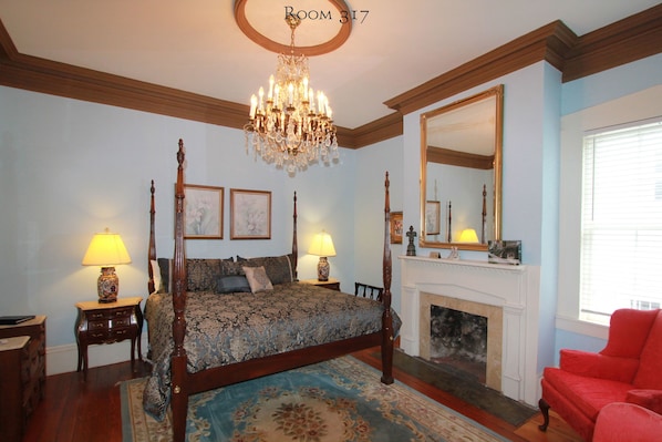A bedroom with a four-poster bed, a chandelier, a fireplace, and a red armchair.