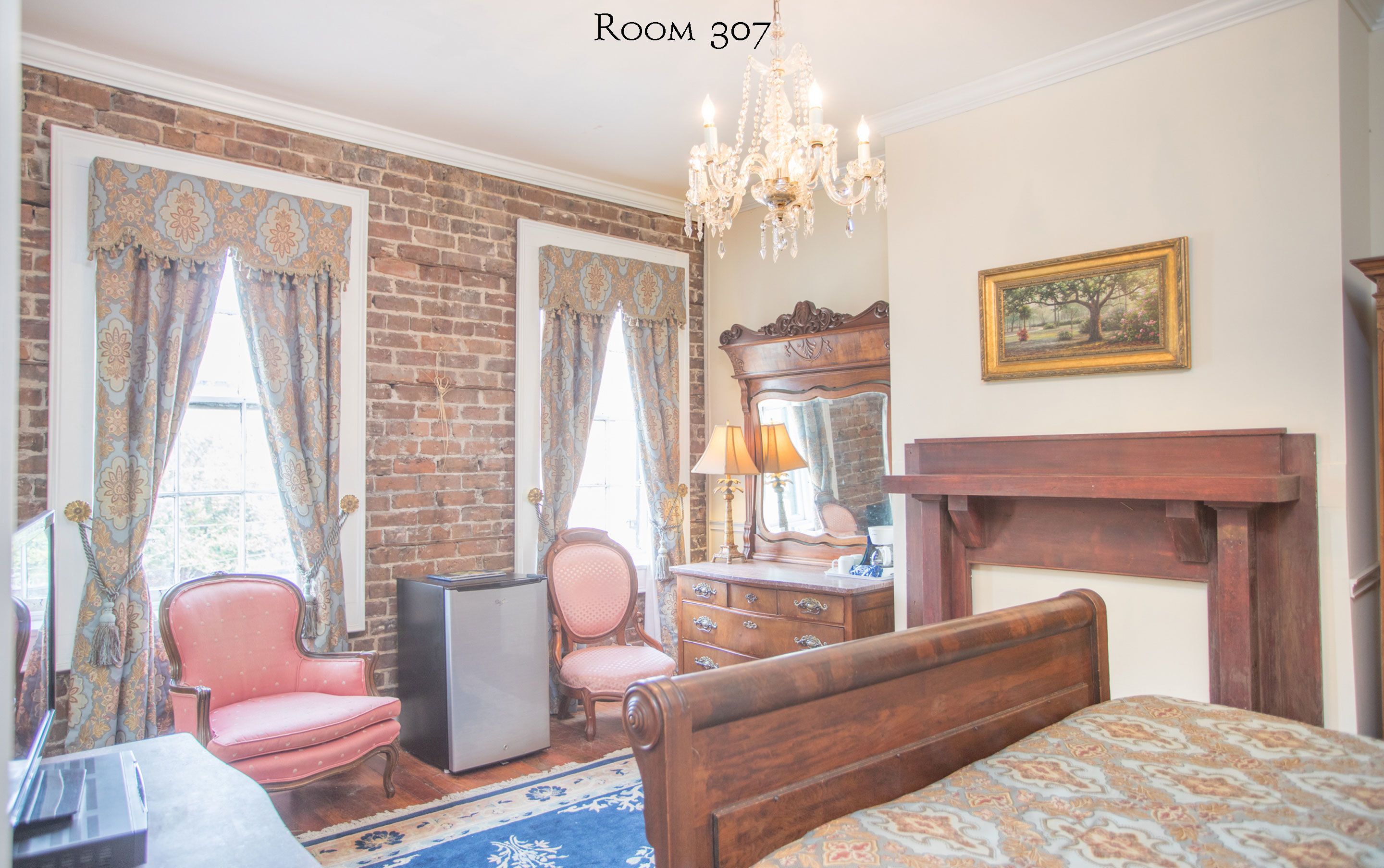 A room with a brick wall, a chandelier, a bed, a dresser, a chair, and a desk with a lamp.