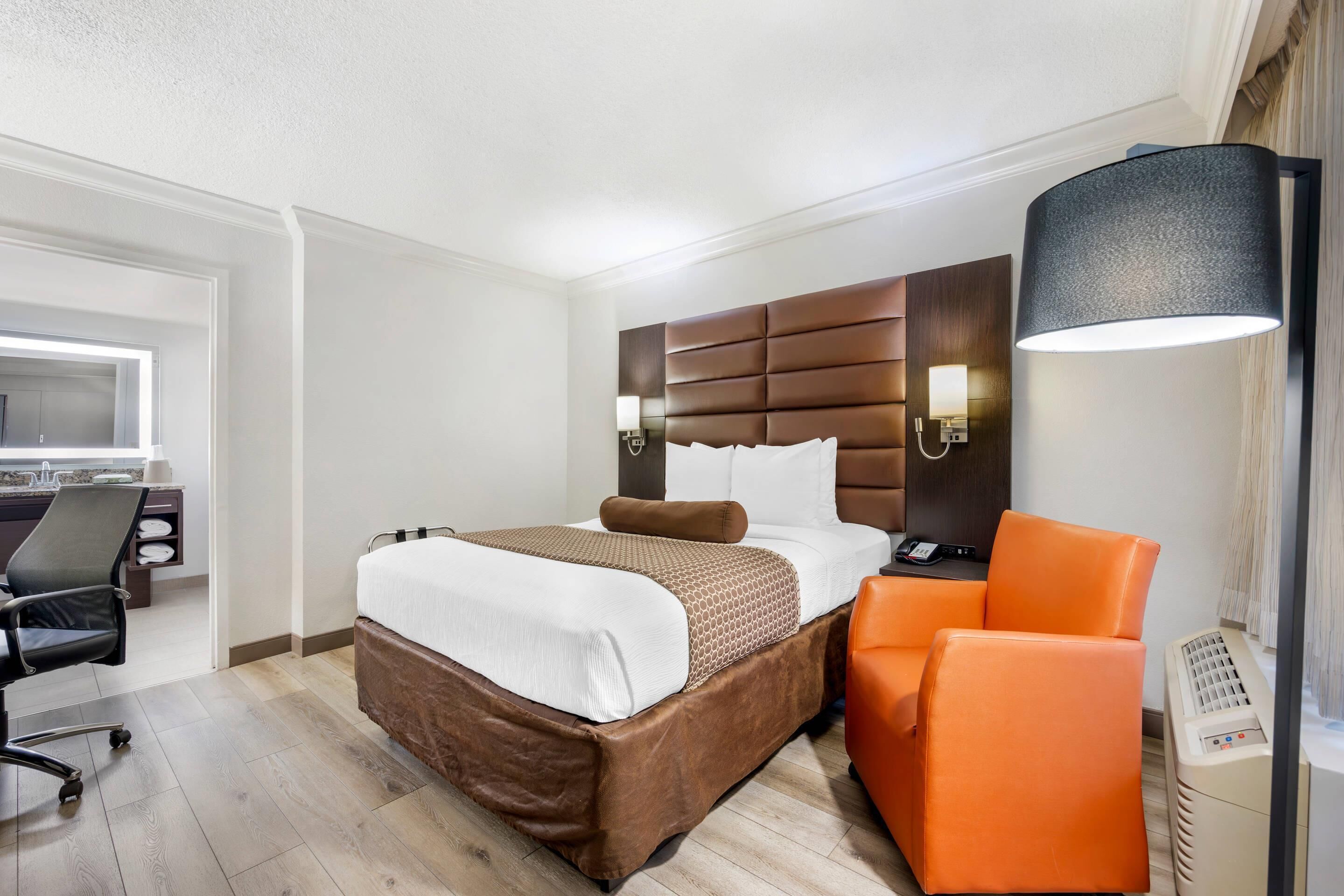 A hotel room with a bed, an orange chair, a desk with a lamp, and a window.