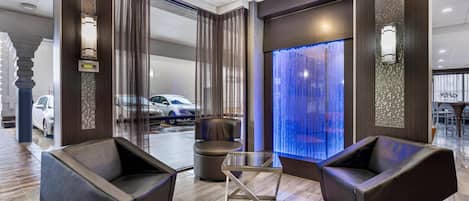 Um lobby moderno com duas poltronas de couro, uma mesa de centro de vidro e um recurso de água com iluminação azul.