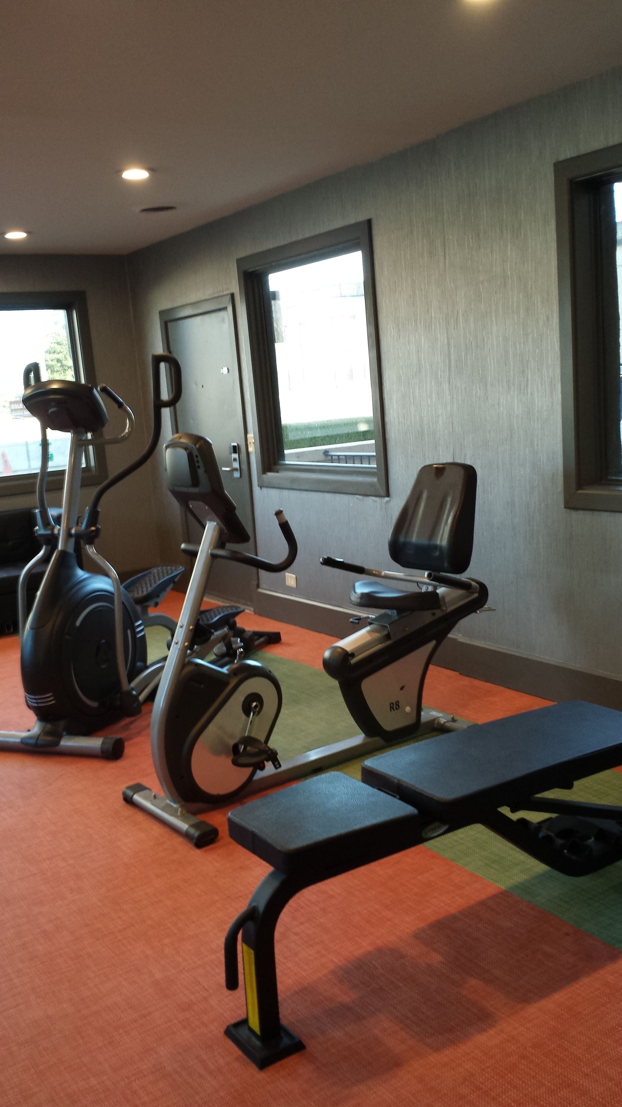 Academia equipada com aparelhos de cardio, um banco e grandes janelas.