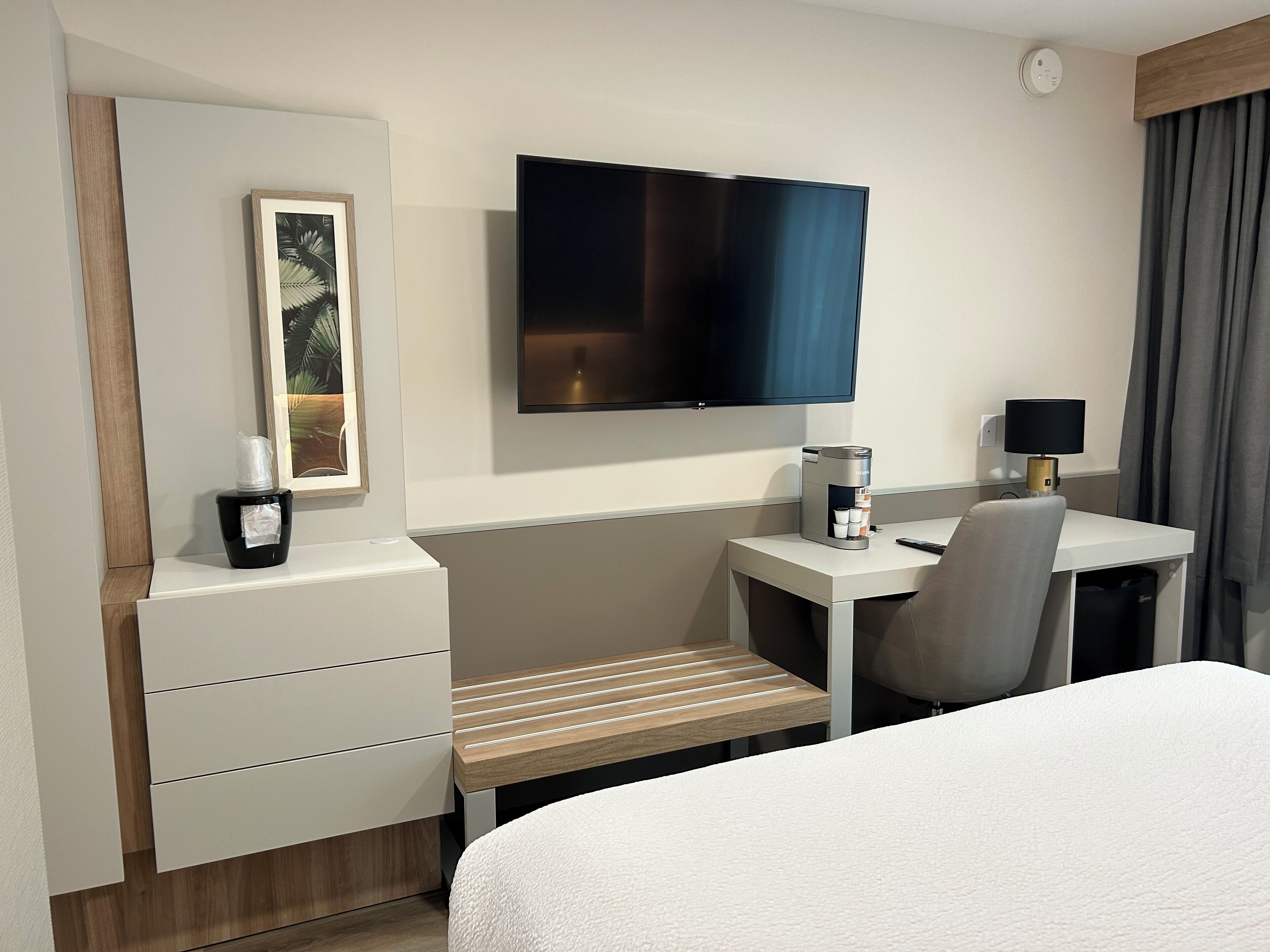 Une chambre d’hôtel comprenant un lit, un bureau avec une chaise, une télévision, une machine à café et une petite table avec une lampe.