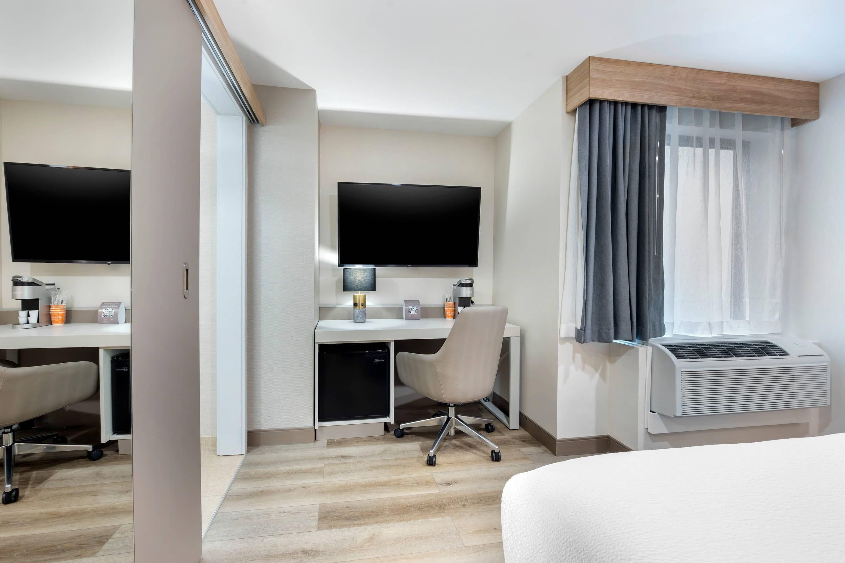 Habitación de hotel con cama, escritorio con silla, televisor de pantalla plana, equipo de aire acondicionado y ventana con cortinas.