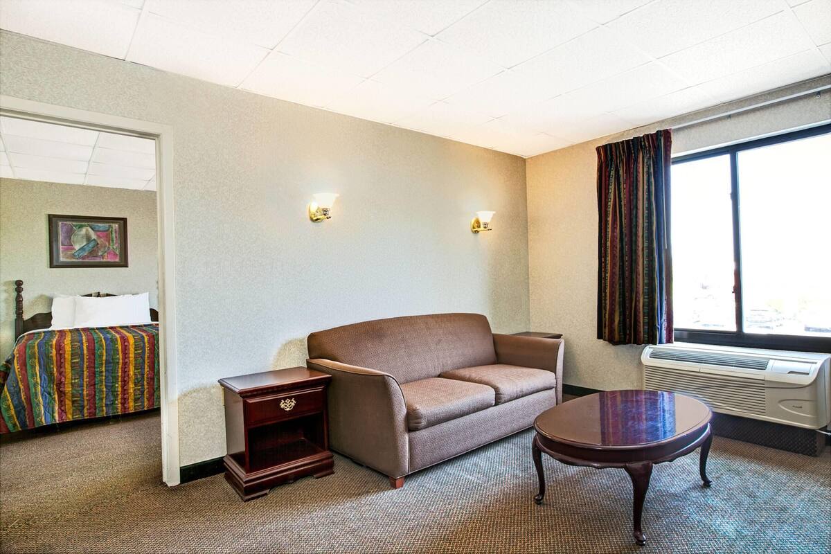 A hotel room with a brown sofa, a small table, a bedside table, a bed with a colorful blanket, two wall sconces, a window with curtains, and an air conditioning unit.