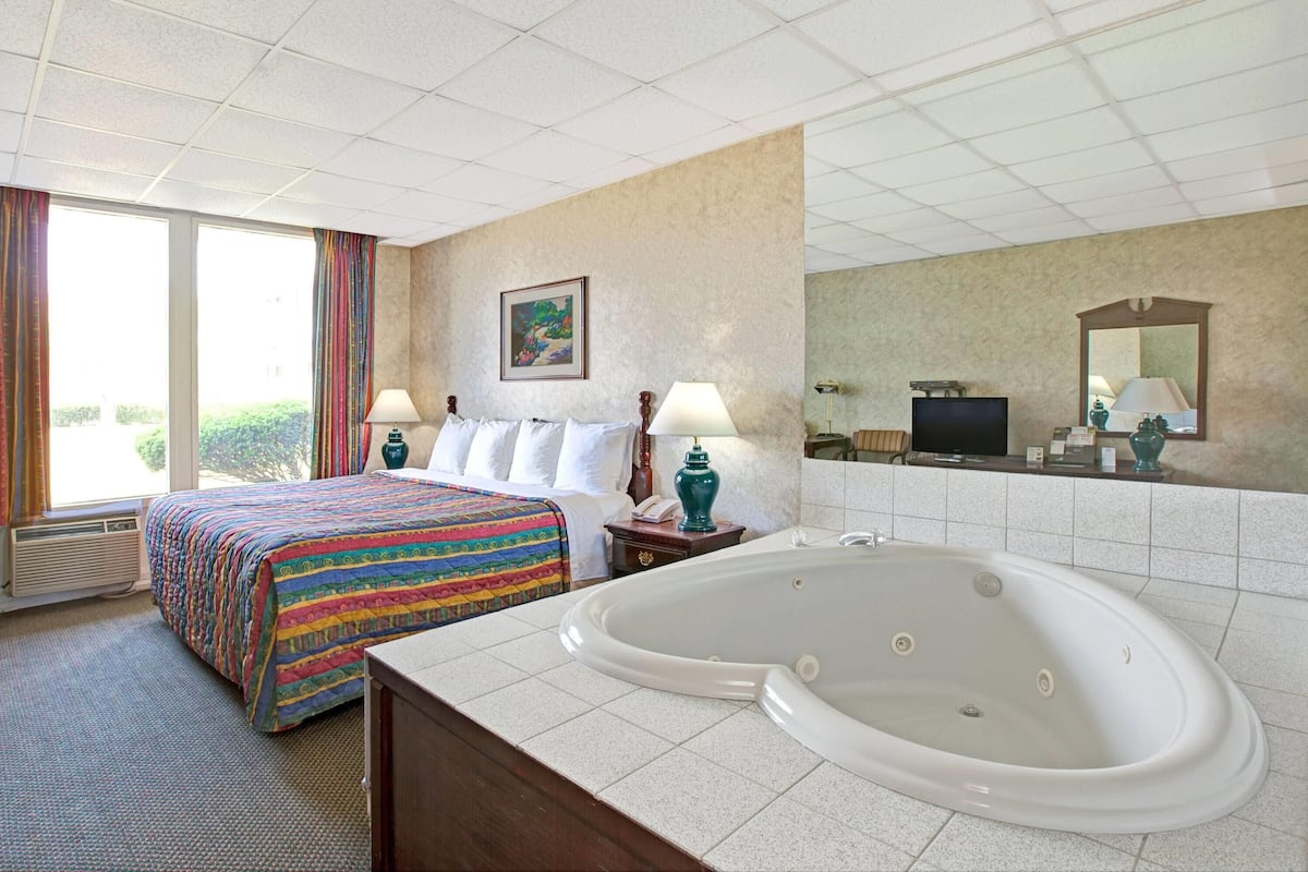 A hotel room with a large bed, a bathtub, a desk with a lamp, and a television.