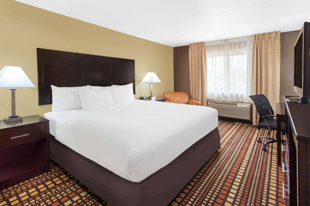 A hotel room with a large bed, two bedside tables with lamps, a desk with a chair, a television, and an air conditioning unit.