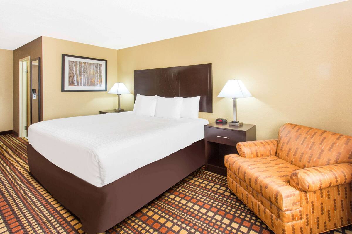 A hotel room with a large bed, two bedside tables with lamps, a patterned armchair, and a framed picture on the wall.