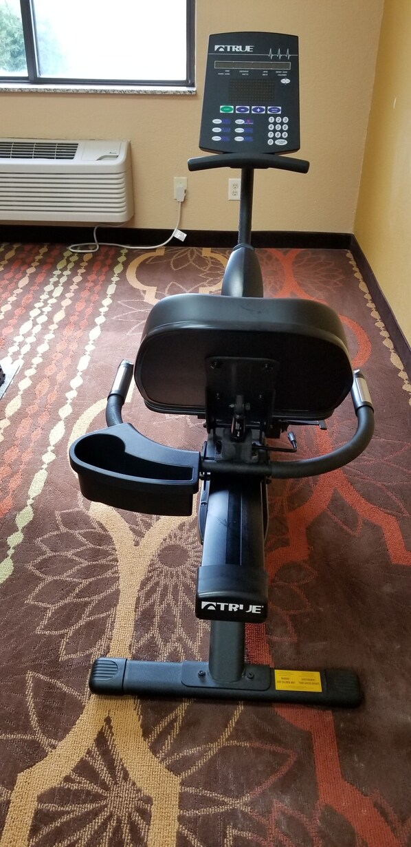 Una bicicleta estática con pantalla digital, ubicada en una habitación con alfombra estampada y una ventana al fondo.