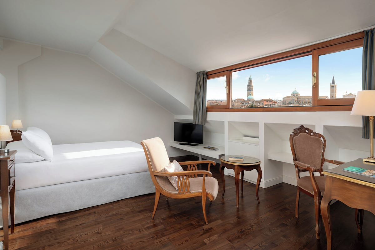 A hotel room with a bed, a television, a wooden chair, a small table, and a view of a cityscape through large windows.