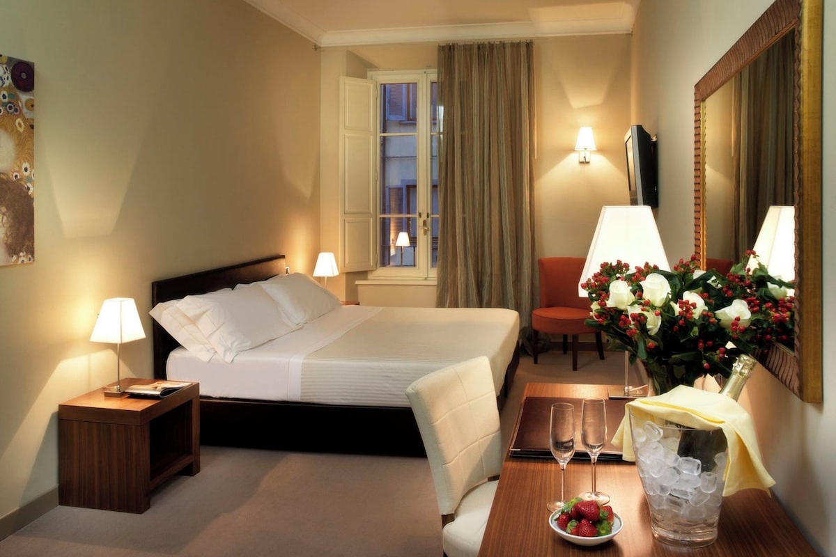 Una habitación de hotel con una cama, lámparas de noche, un escritorio con un florero y un espejo, además de una ventana con cortinas.