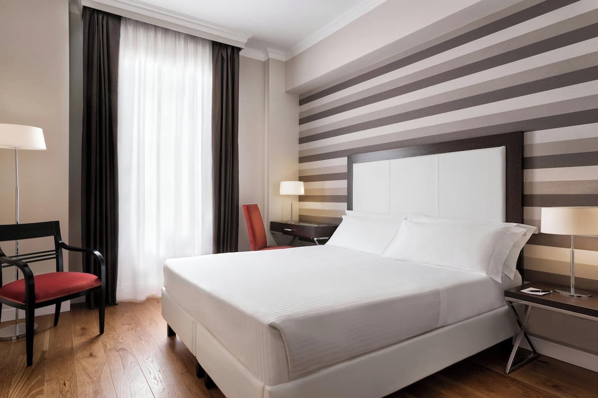 A hotel room with a large bed, a desk, a chair, a lamp, and a window with curtains.