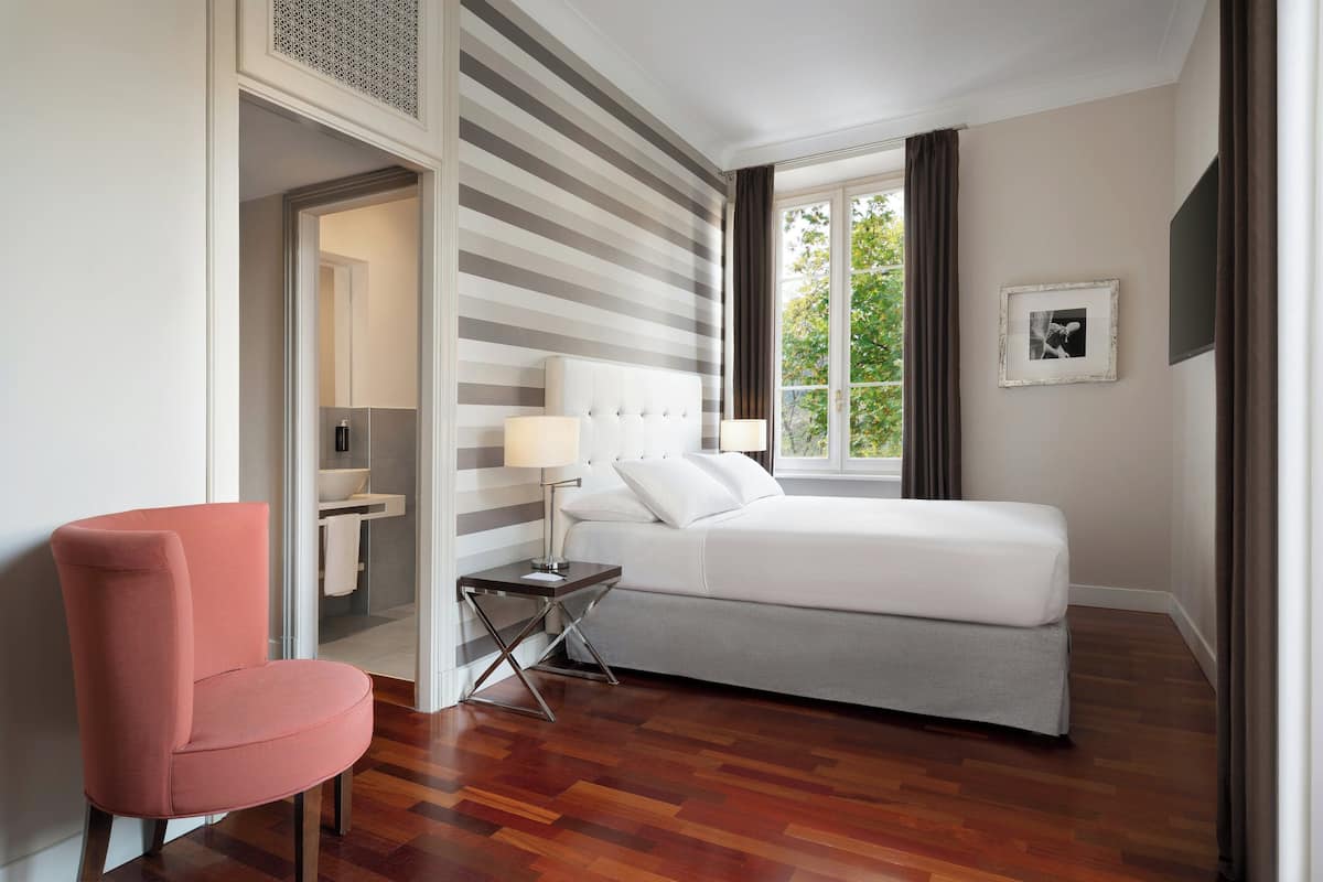 A modern hotel room with a bed, a chair, a side table, a window with curtains, and a bathroom visible through an open door.