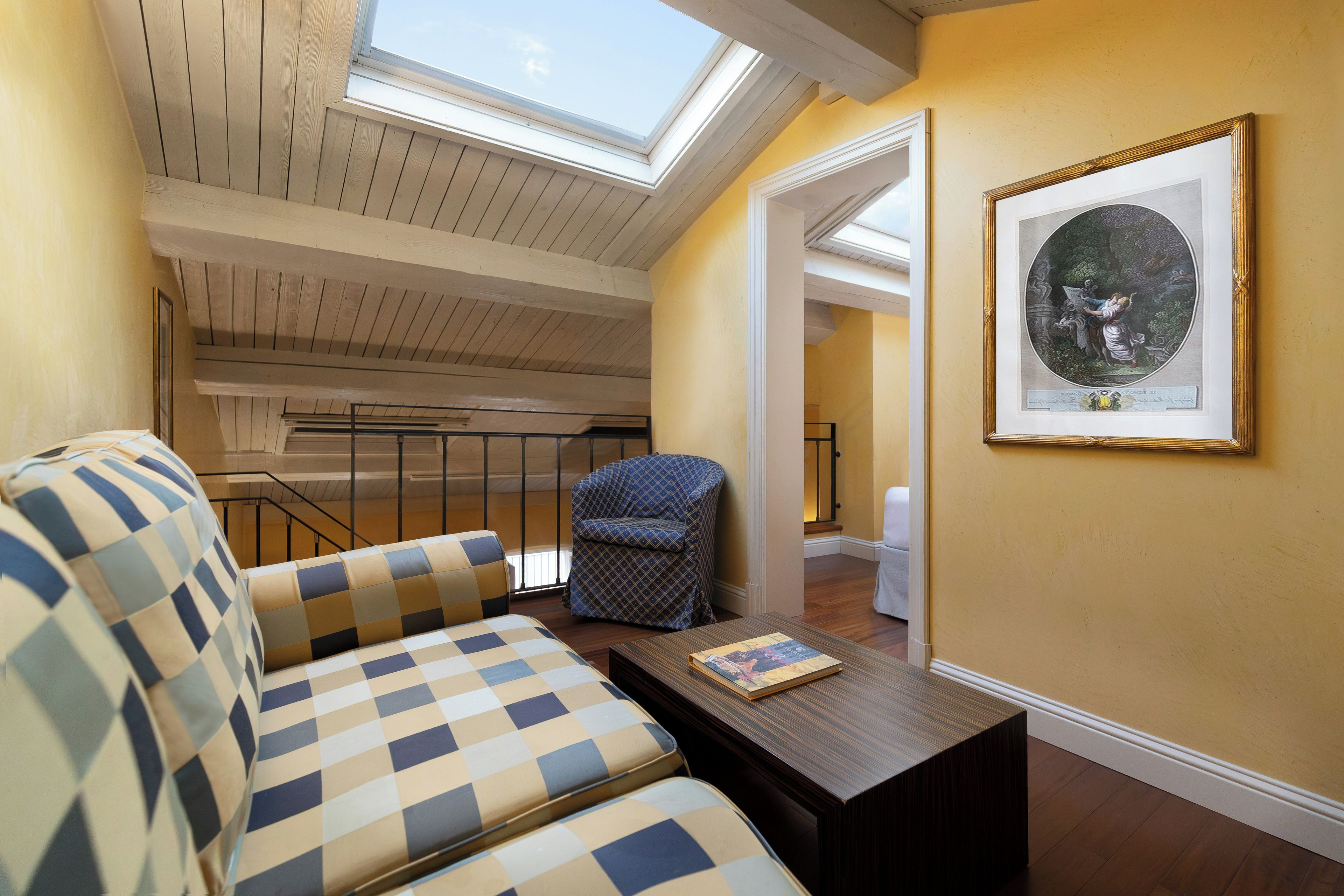 Quarto com sofá xadrez, poltrona estampada, mesa de centro de madeira, obra de arte emoldurada e um claraboia.