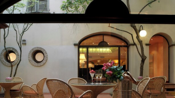 An outdoor seating area with wicker furniture, a table with a vase of flowers, and a view of the interior through an arched doorway. - Sina Maria Luigia (Parma)
