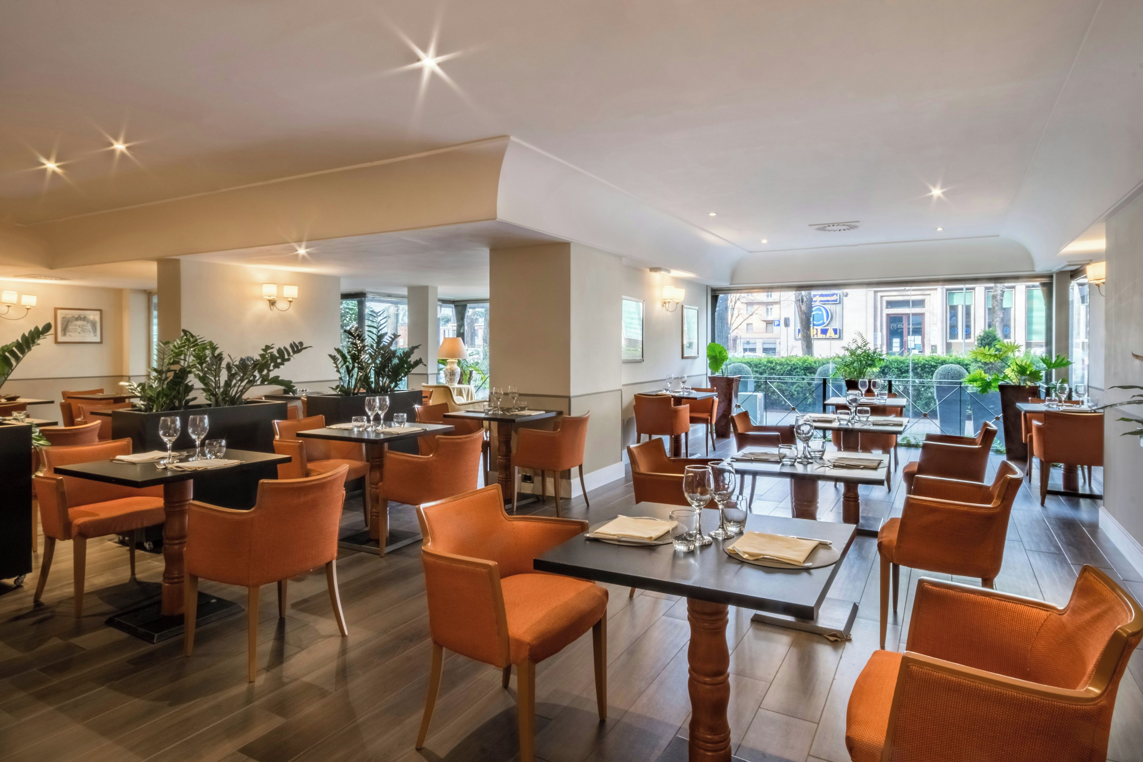 A spacious restaurant with multiple tables and chairs, large windows, and indoor plants.