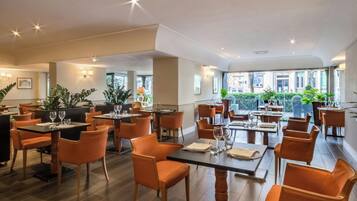 A spacious restaurant with multiple tables and chairs, large windows, and indoor plants.