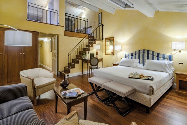 A hotel room with a bed, a chair, a table, a lamp, a nightstand, a mirror, and a staircase.