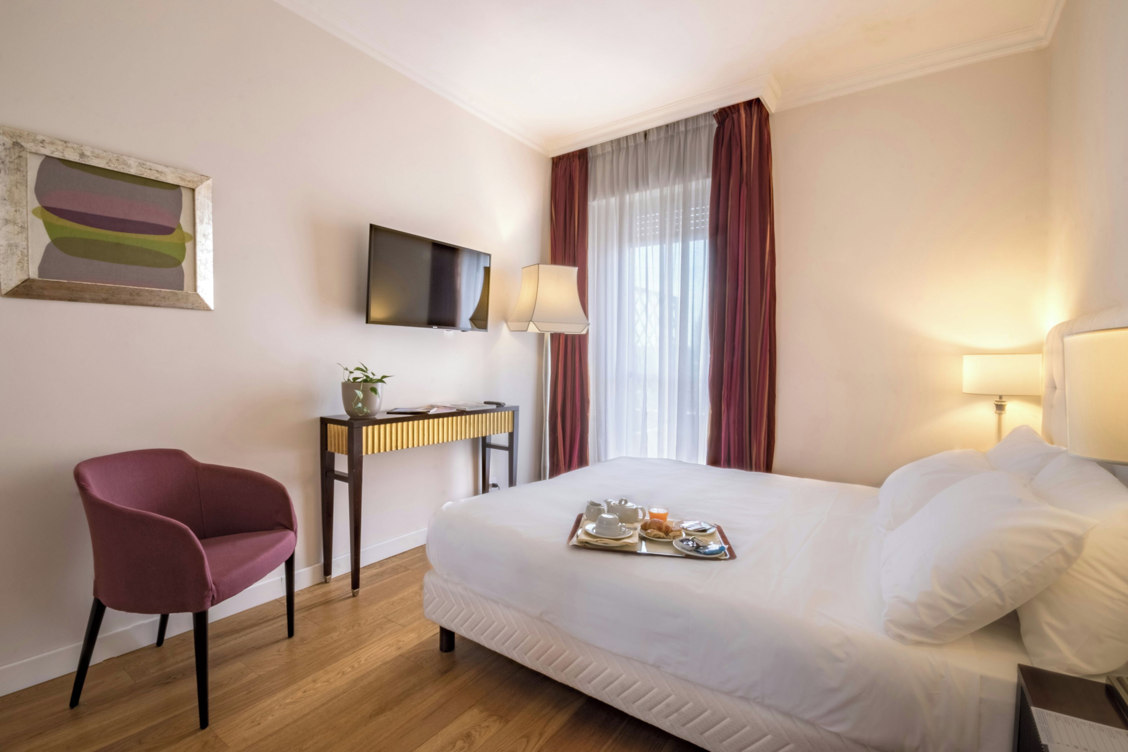 A hotel room with a bed, a desk with a computer, a chair, a TV, and a window with curtains.