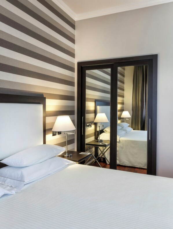 A hotel room with a bed, bedside lamps, a mirror, and a striped wall.